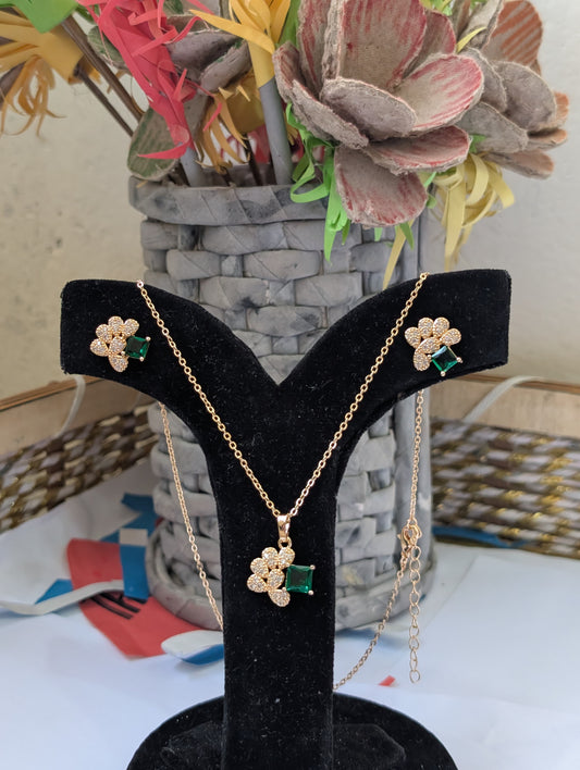 Gold necklace with floral pendant and green gemstones on a black display stand with flowers in the background.
